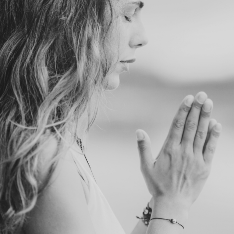 Frau mit langen, welligen Haaren in meditativer Pose mit gefalteten Händen.